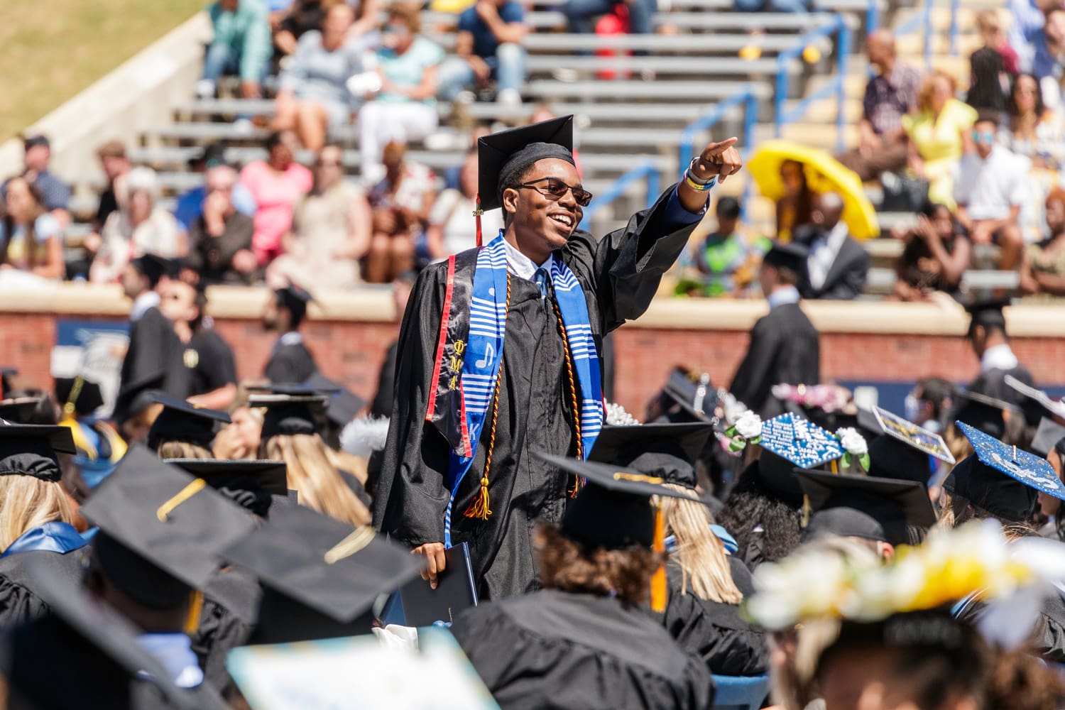 student in graduation cap and gown standing up pointing to the stands 